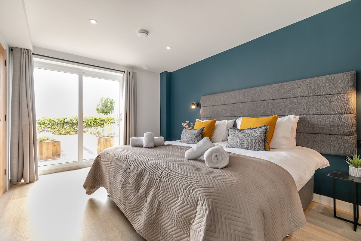 A modern bedroom with a large bed, a view to the outdoors, and a small side table with a plant.