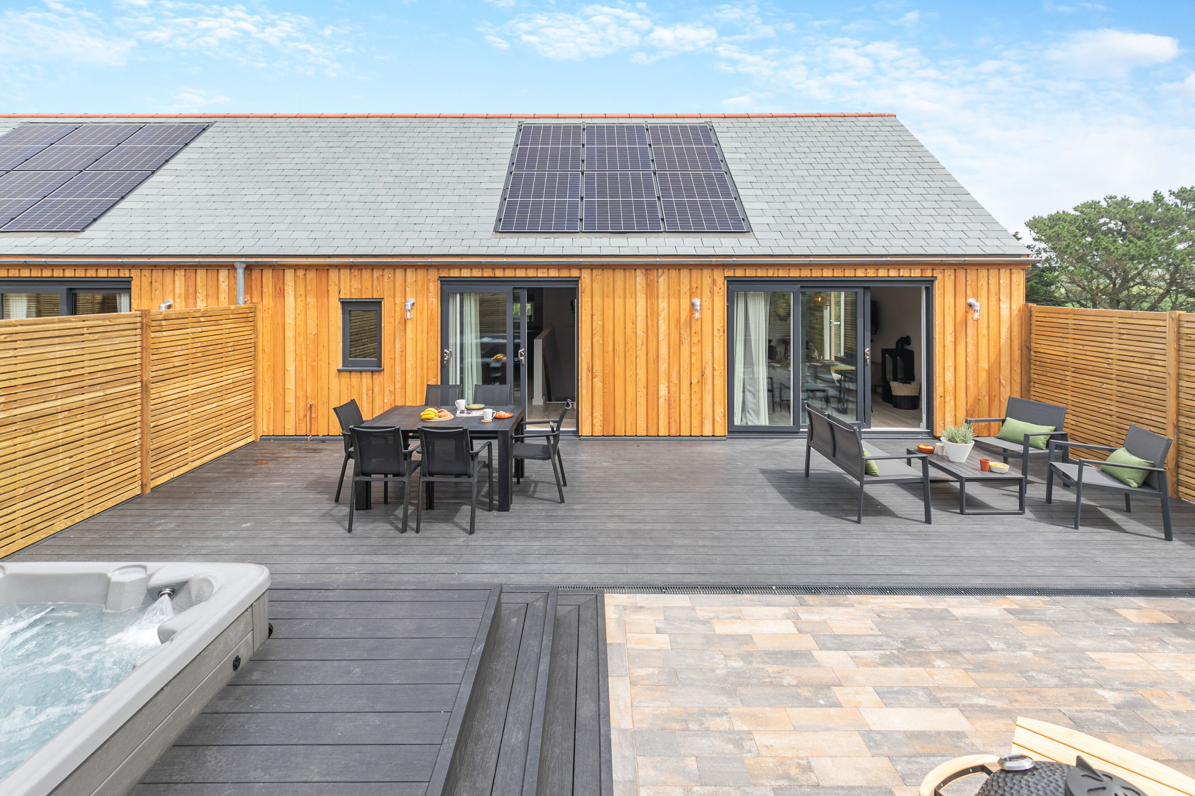 Ein Gebäude aus Holz mit einem geneigten Dach, Solaranlagen und einer Terrasse mit Gartenmöbeln.