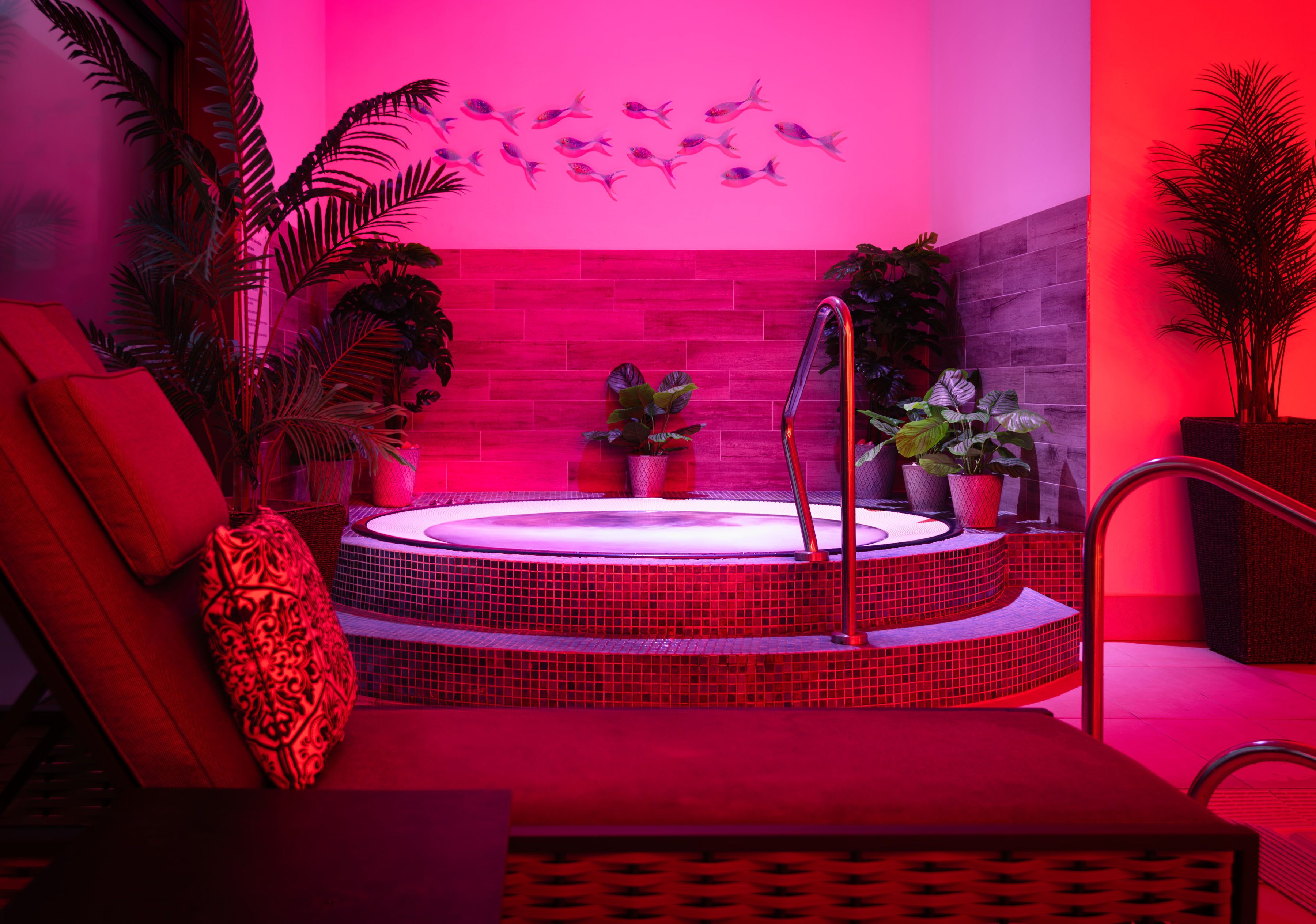 Ein Wellnessbereich mit Whirlpool, roter und pinkfarbener Beleuchtung und diversen Pflanzen.
