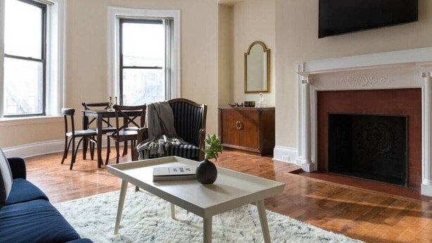 A living room with a fireplace, a television mounted above it, a dining area with a table and chairs, and a large window providing natural light. - Beautiful 1BR near Fenway Park + Kenmore! (Boston)