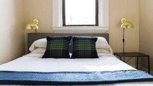 A bed with white linens, two plaid pillows, and a blue throw blanket. There are two bedside lamps and a small table beside the bed. - Beautiful 1BR near Fenway Park + Kenmore! (Boston)