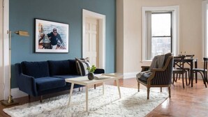 A living room with a dark blue sofa, a white coffee table, a striped armchair, and a dining table with chairs. - Beautiful 1BR near Fenway Park + Kenmore! (Boston)