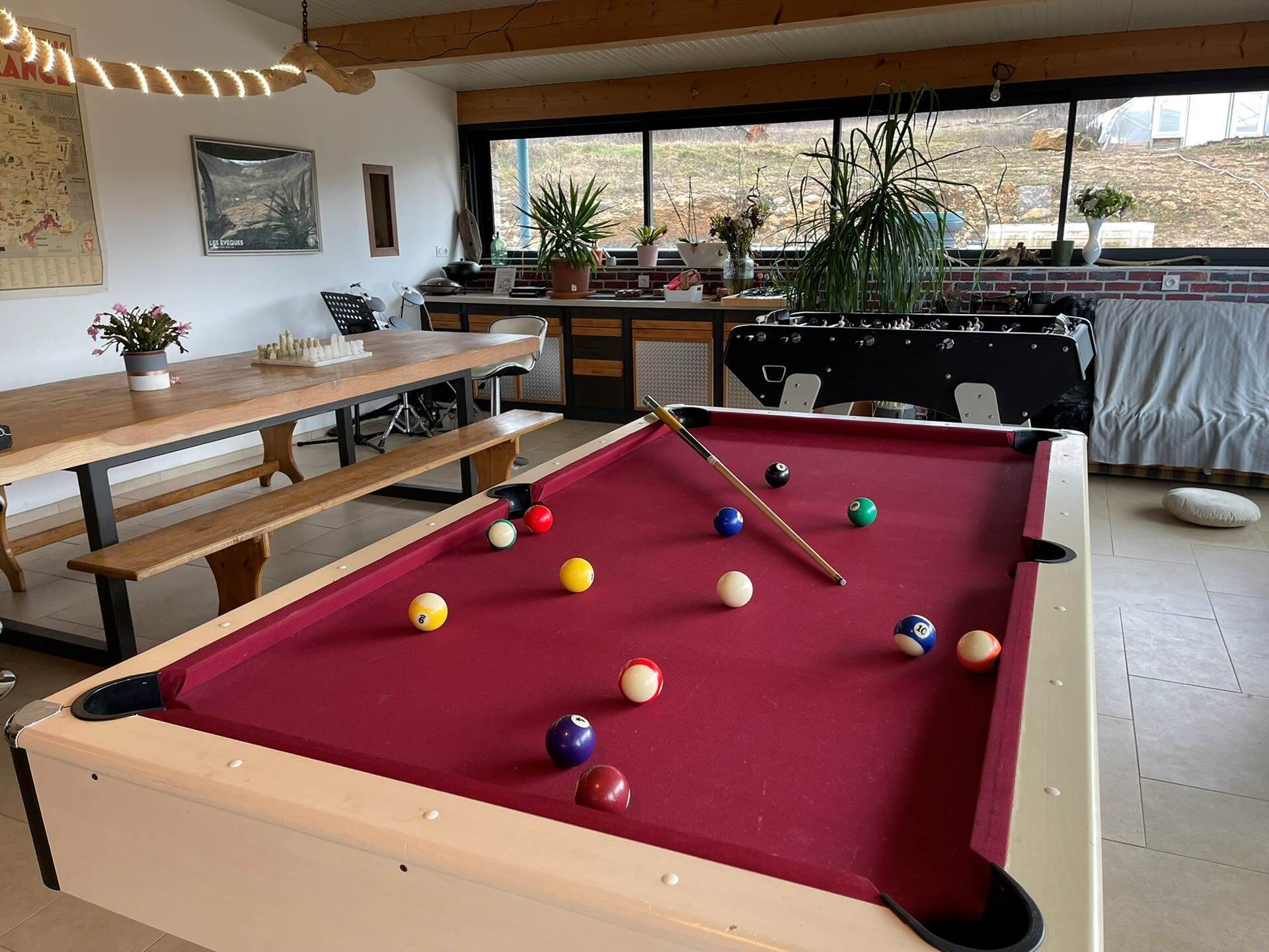 Un tavolo da biliardo con le palle, un tavolo in legno e sedie in una stanza con una grande finestra.