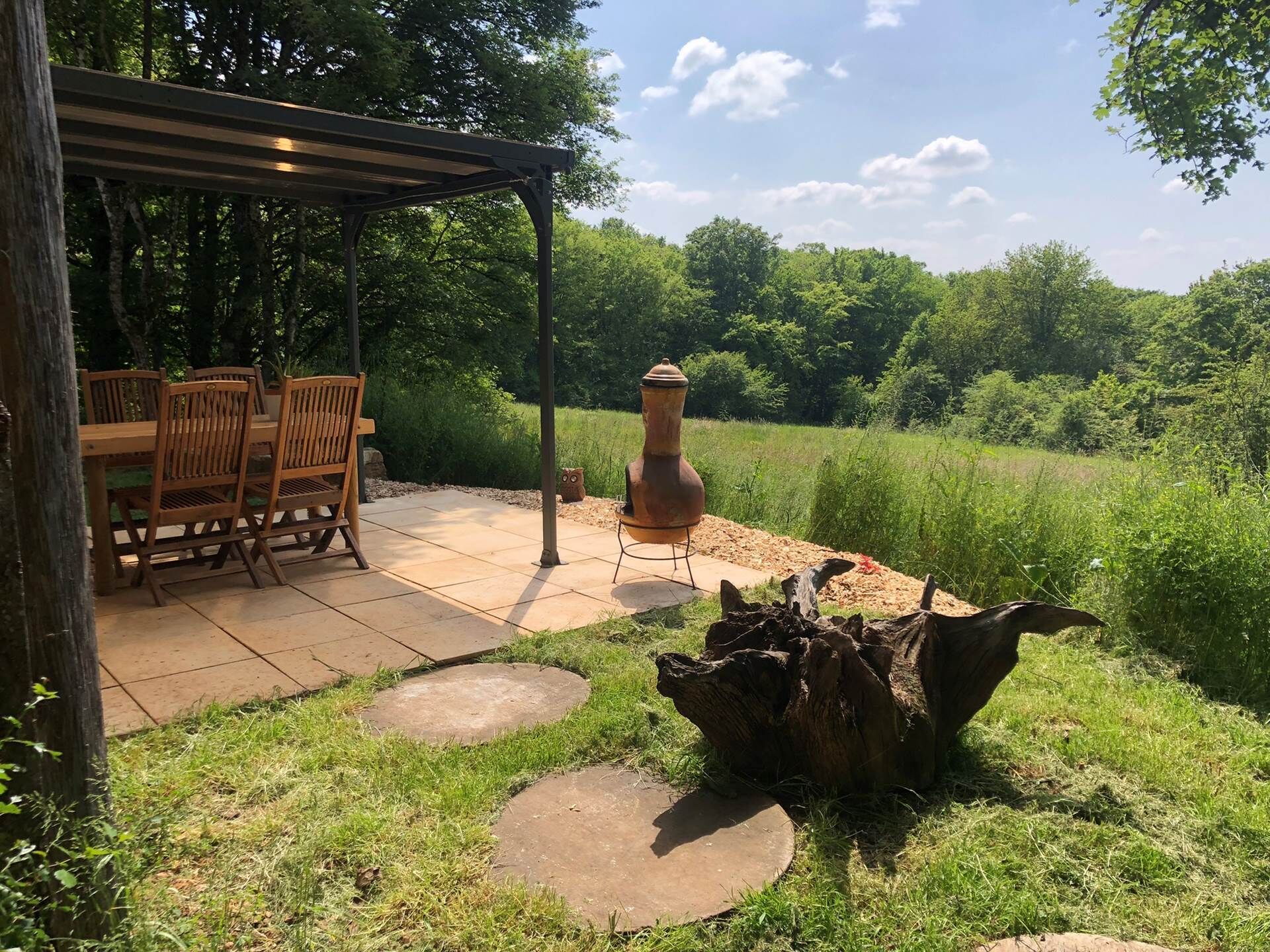 An outdoor patio with a wooden table and chairs, a rustic fire pit, and a pergola.