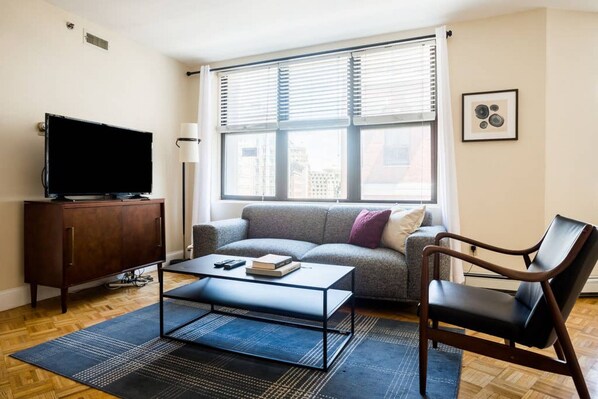A living room with a sofa, a coffee table, a TV, a chair, and a window with blinds. - Walking distance to Beacon Hill, Back Bay + all tourist spots.  Great for work trip too, MBTA nearby (Boston)