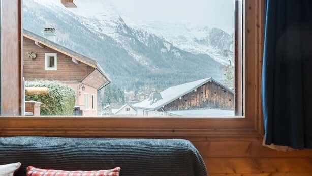 Una habitación con paredes de madera, una ventana que ofrece vistas a montañas nevadas y edificios, y una cama con un cojín escocés.