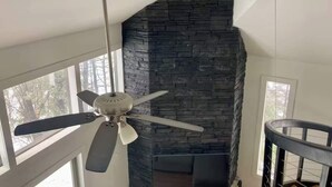 A room with a stone fireplace, a ceiling fan, and large windows. - Newly Renovated 3 Bedroom! Walk to Bars and Restaurants (Killington)