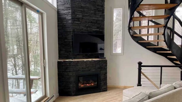 A living room with a stone fireplace, a spiral staircase, and a large window with a view of trees. - Newly Renovated 3 Bedroom! Walk to Bars and Restaurants (Killington)