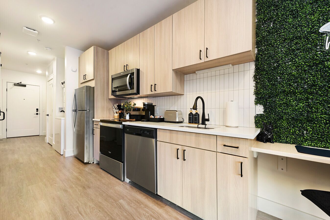 A modern kitchen with light wood cabinets, stainless steel appliances, a microwave, and a green wall feature.