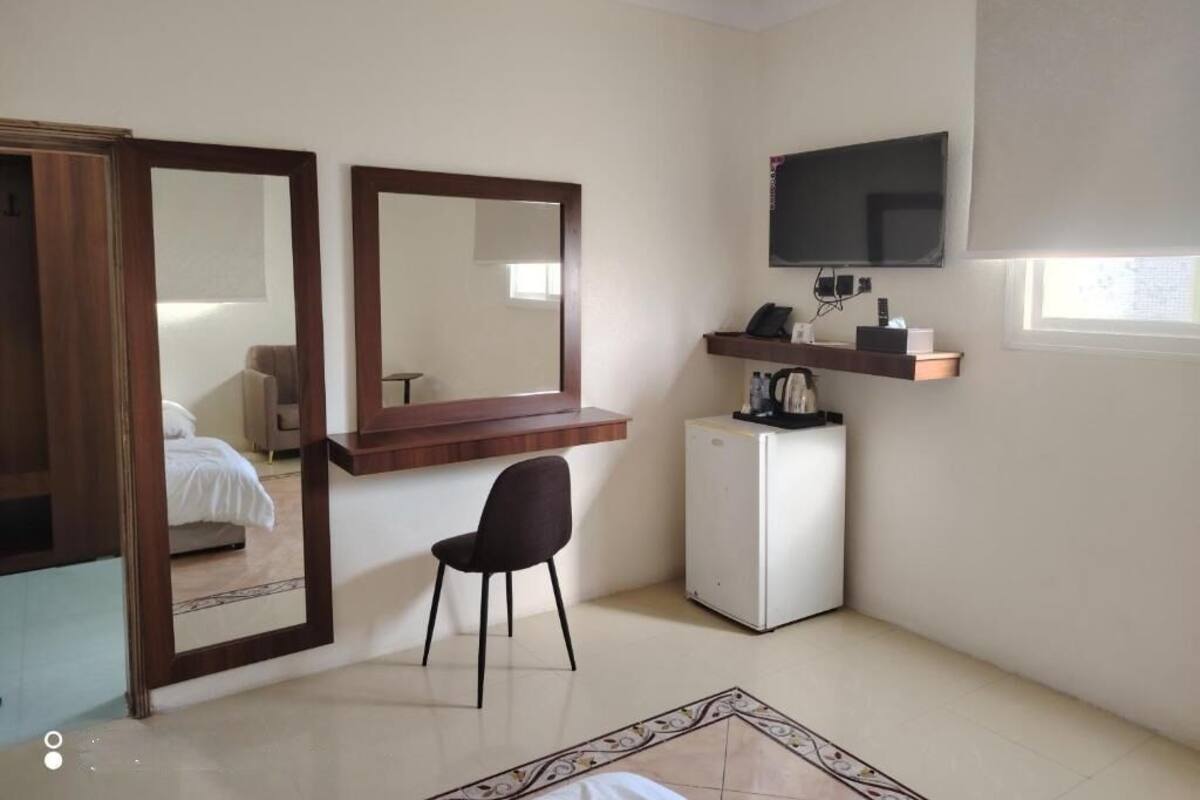 A compact hotel room with a bed, a desk with a chair, a television mounted on the wall, a small refrigerator, and a window with a blind.