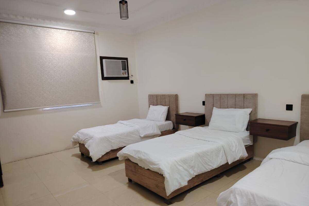 A hotel room with three single beds, a bedside table with a lamp, a wall-mounted air conditioner, and a window with blinds.