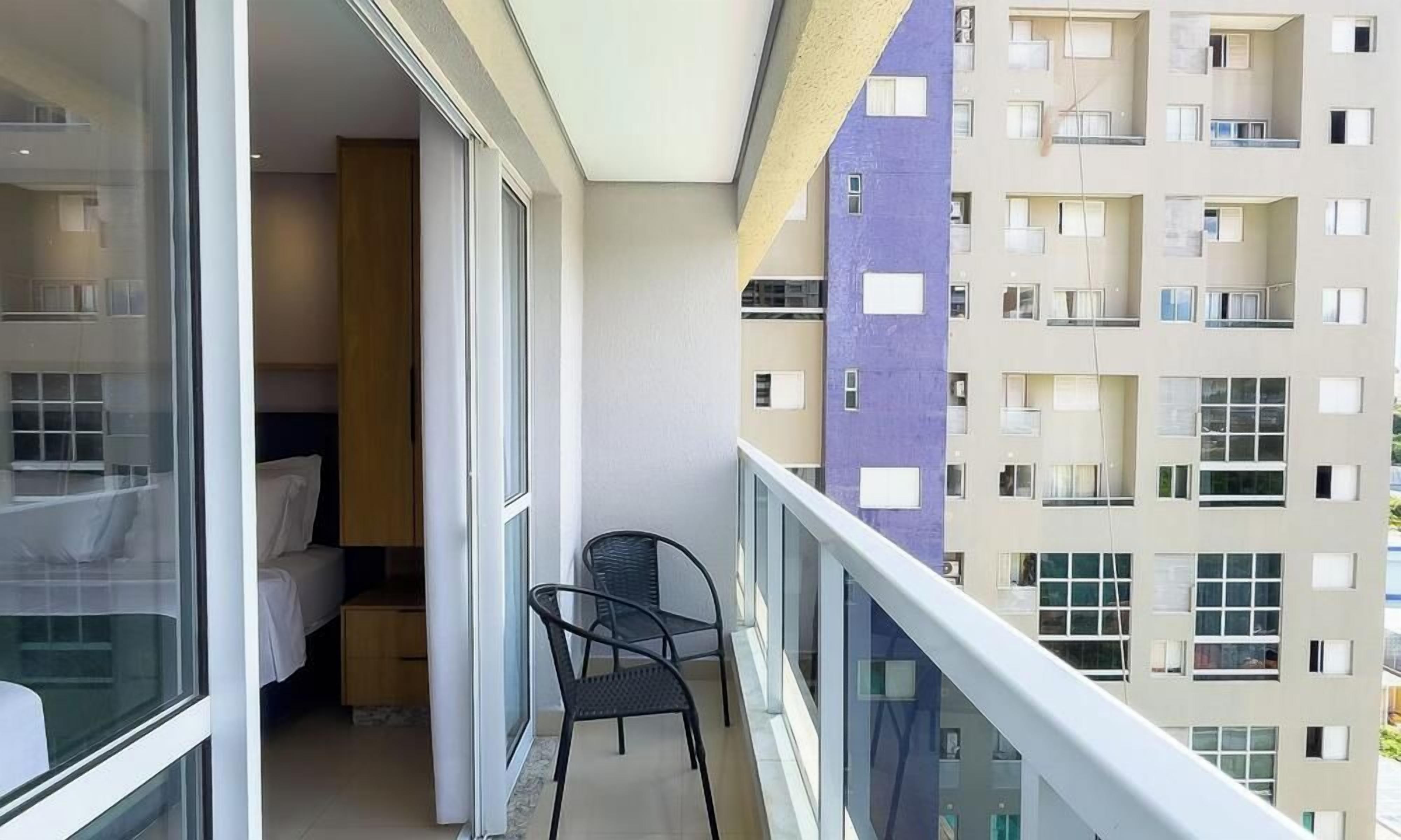 A balcony with a bed, a chair, and a view of a multi-story building.