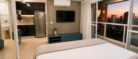 A modern hotel room with a bed, a TV, a kitchenette, and a city view.