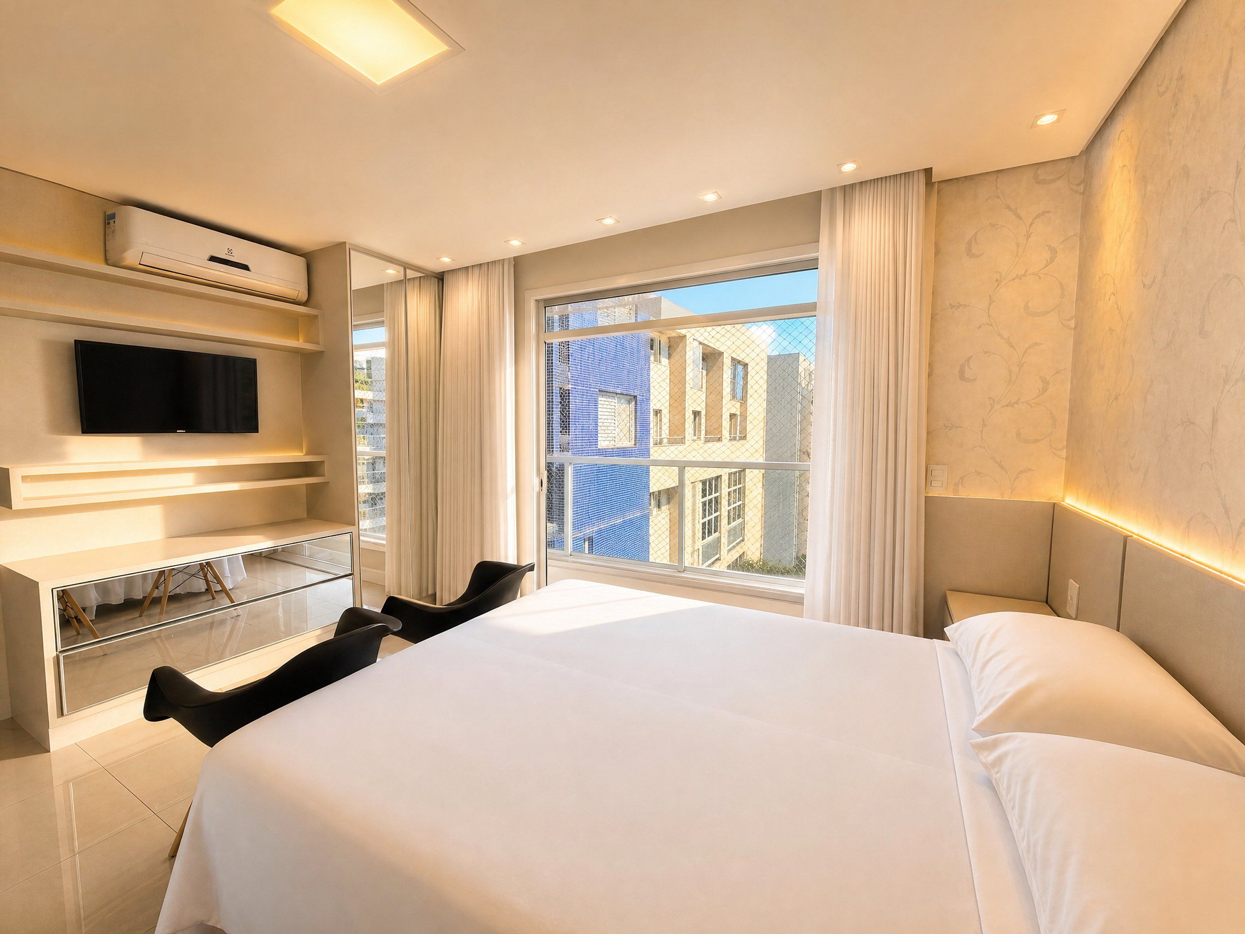 A hotel room with a large bed, a flat-screen TV mounted on the wall, a glass desk, and a view of buildings through the window.