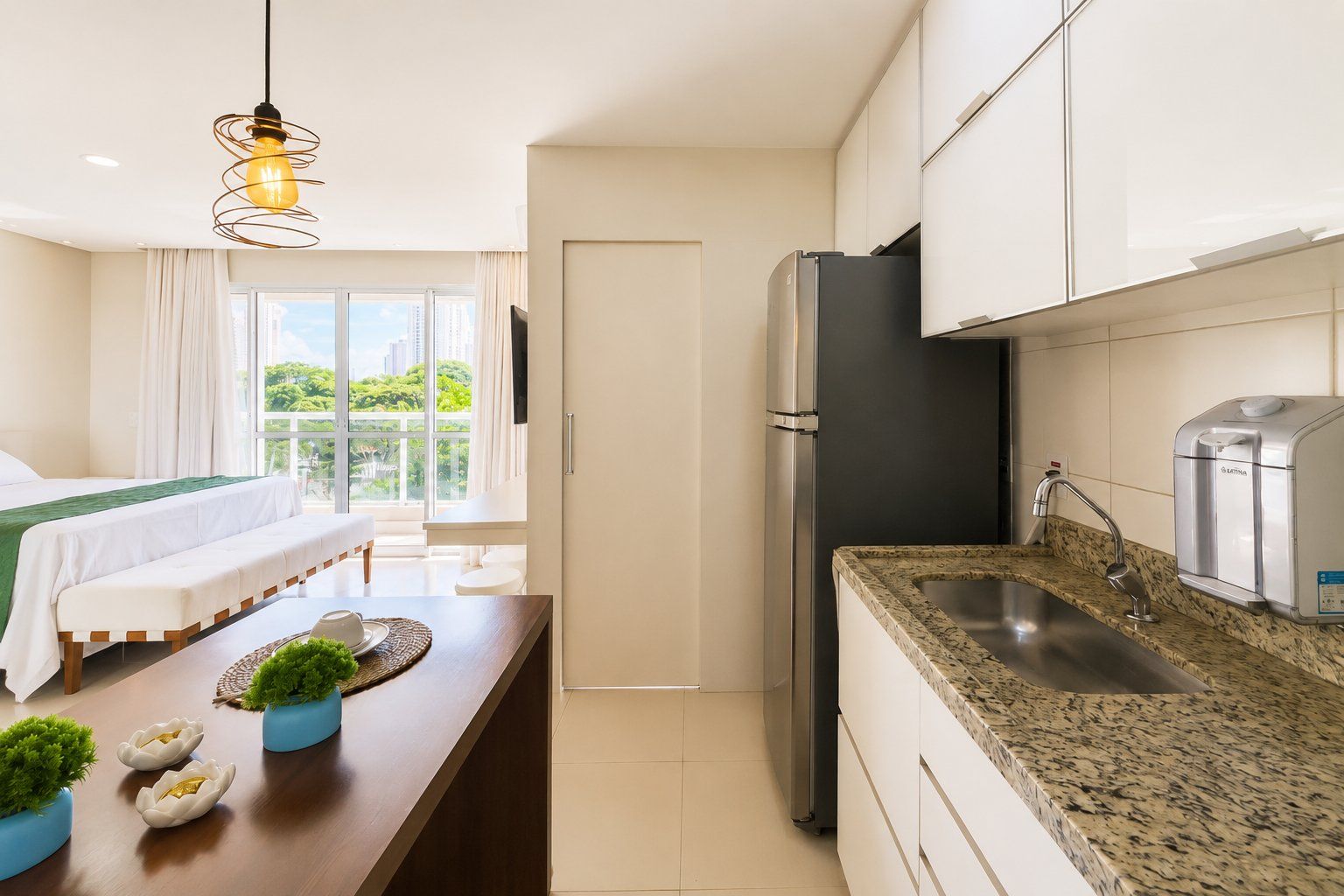A modern hotel room with a bed, a dining area, a kitchen with a refrigerator, and a view of greenery.