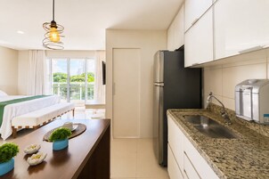 Quarto de hotel moderno com cama, área de refeições, cozinha equipada com frigorífico e vista para área verde.