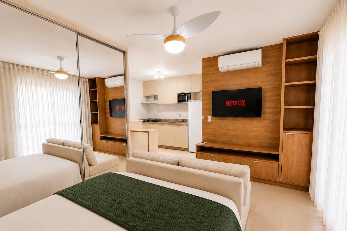 A modern hotel room with a bed, a sofa, a TV, a kitchenette, and a ceiling fan.