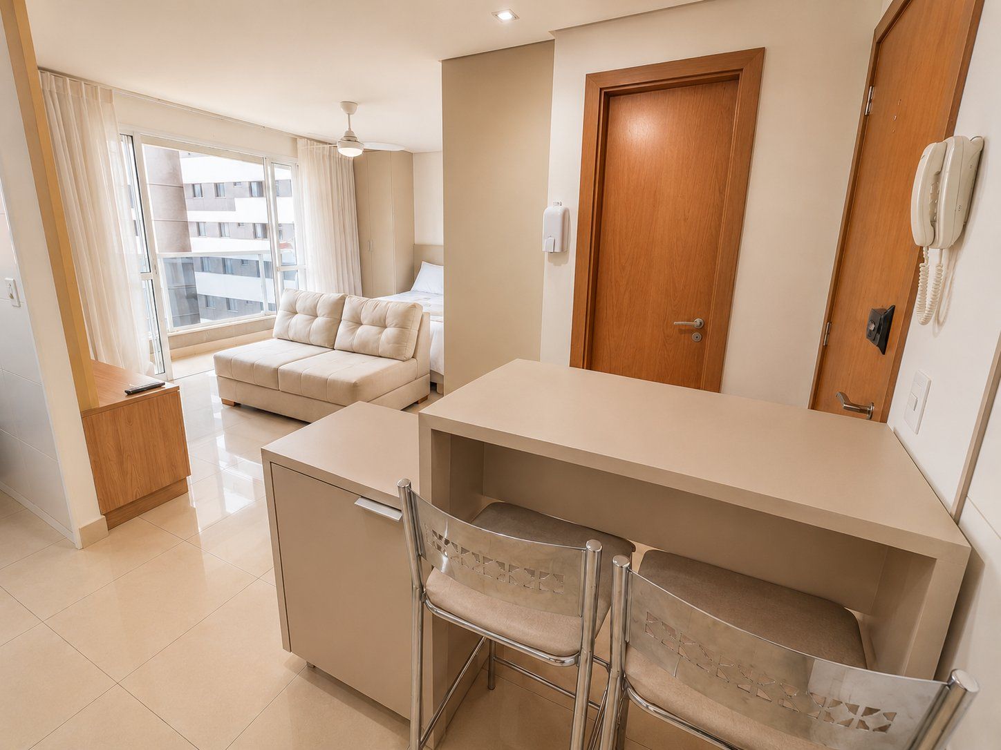 A compact hotel room with a bed, a sofa, a desk with chairs, a wooden door, and a phone.