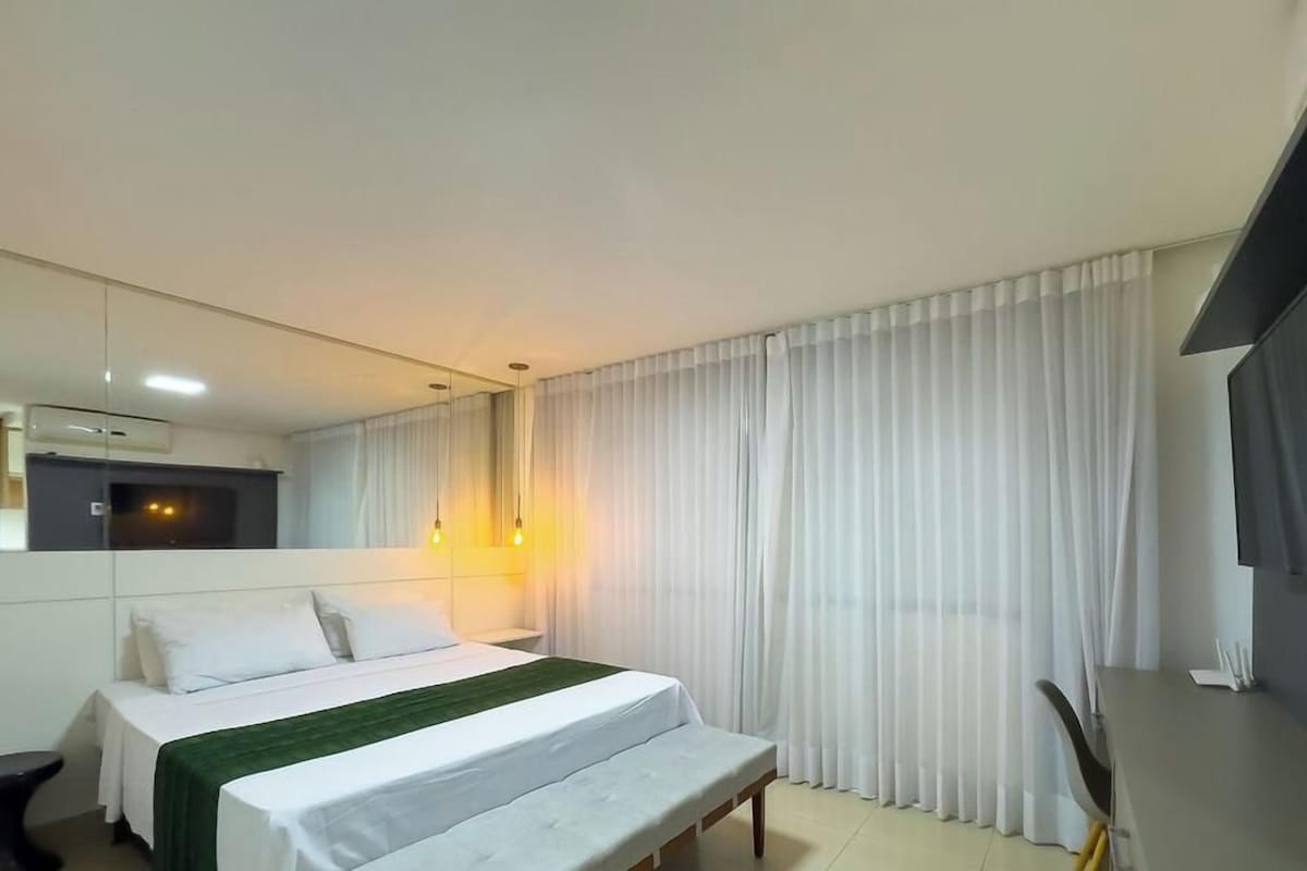 A hotel room with a bed, a bench, a TV, and a large window with curtains.