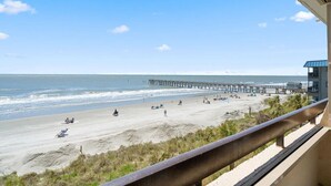 Uitzicht op een strand met een pier die zich uitstrekt over de oceaan, zandstrand en een gebouw met een balkon.