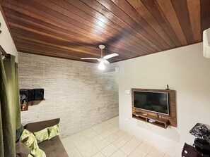 Ein Wohnzimmer mit Holzboden, einem an der Wand befestigten Flachbildfernseher, einem Deckenventilator und einer Wand im Ziegelstein-Design.