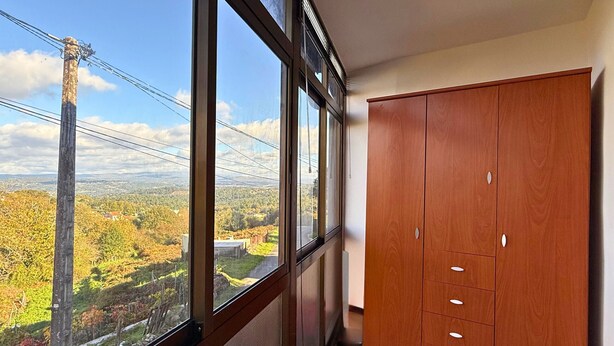 Een kamer met grote ramen, met uitzicht op een landschap met bomen en een elektriciteitsleiding. Er staat een houten garderobe met meerdere lades.