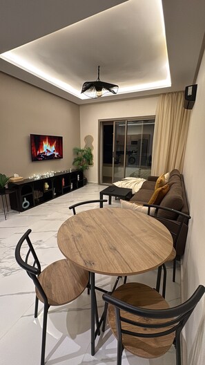 A modern living room with a round wooden table and chairs, a flat-screen TV mounted on the wall, and a ceiling light fixture. - Comfort & Sun : Pool view, Beach 400m (Âïn-Harrouda)