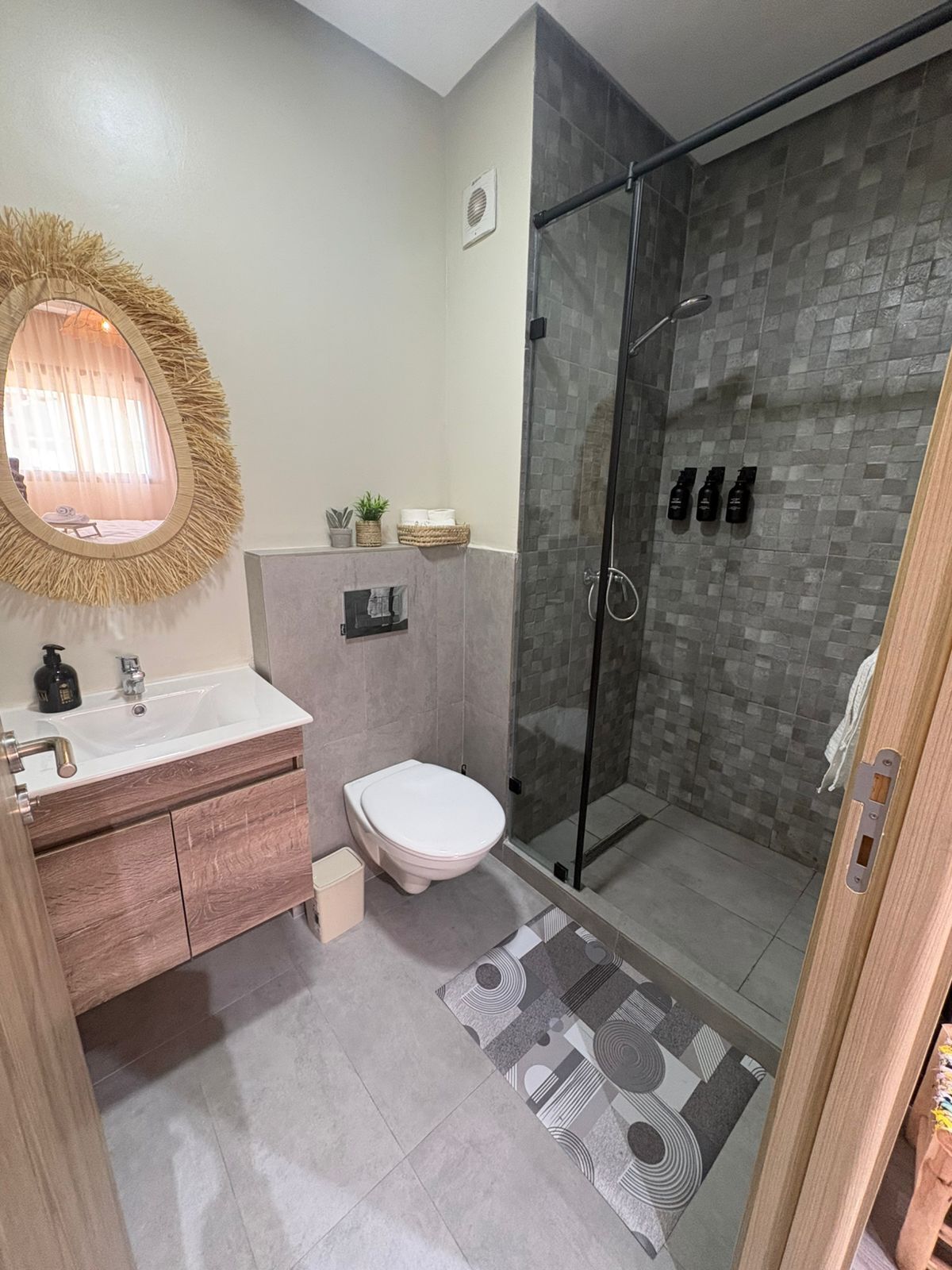 Ein modernes Badezimmer mit Duschkabine aus Glas, einem runden Spiegel mit strohähnlichem Rahmen, einer weißen Toilette und einem Waschtisch mit Holdekor.