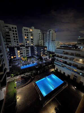 Ett modernt bostadskomplex med flera höga byggnader, en central innergård med en swimmingpool och väl upplysta utomhusområden.