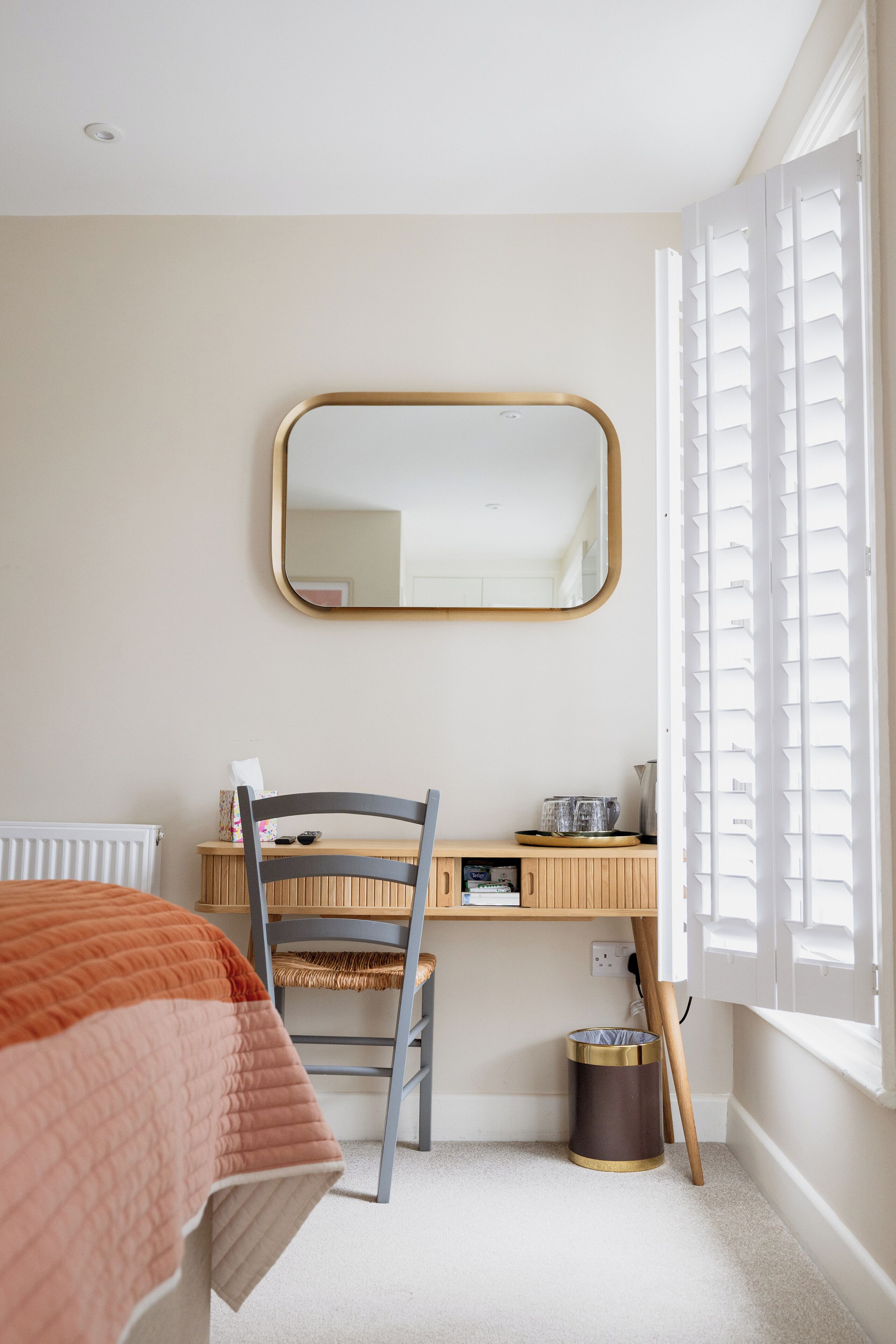 Un dormitorio con cama, escritorio, silla, espejo, radiador, ventana con persianas y cubo de basura.