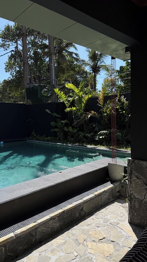 Ein Pool mit einer Steindeck, umgeben von tropischen Pflanzen und blauem Himmel.