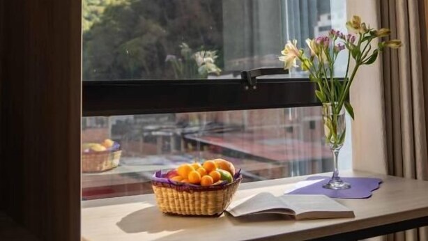 Una habitación con vistas a montañas a través de una ventana, una mesa con una cesta de fruta, un florero con flores y un libro abierto.