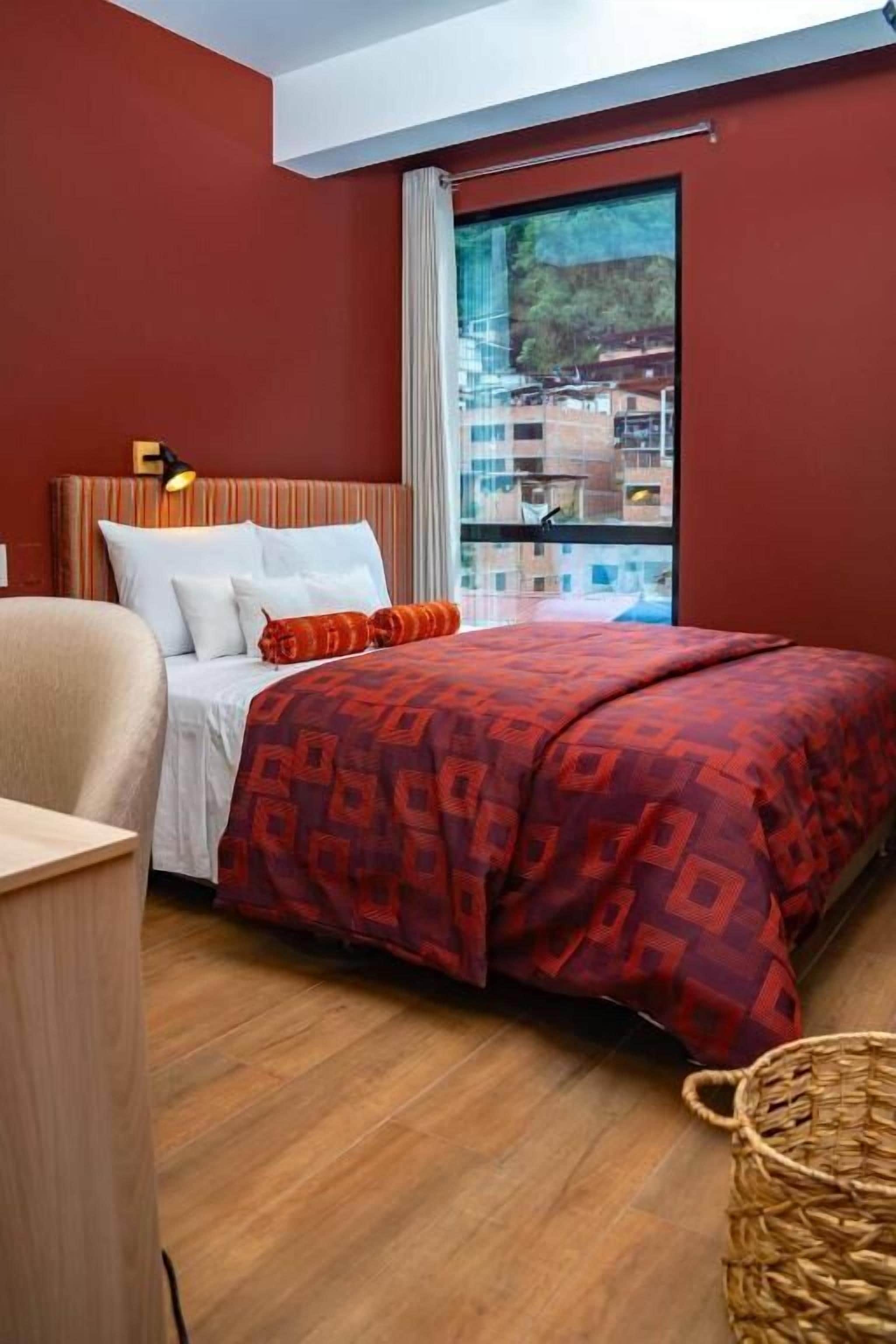 Habitació d'hotel amb un llit, una cadira, una finestra i vistes a edificis, i un sòl de fusta.