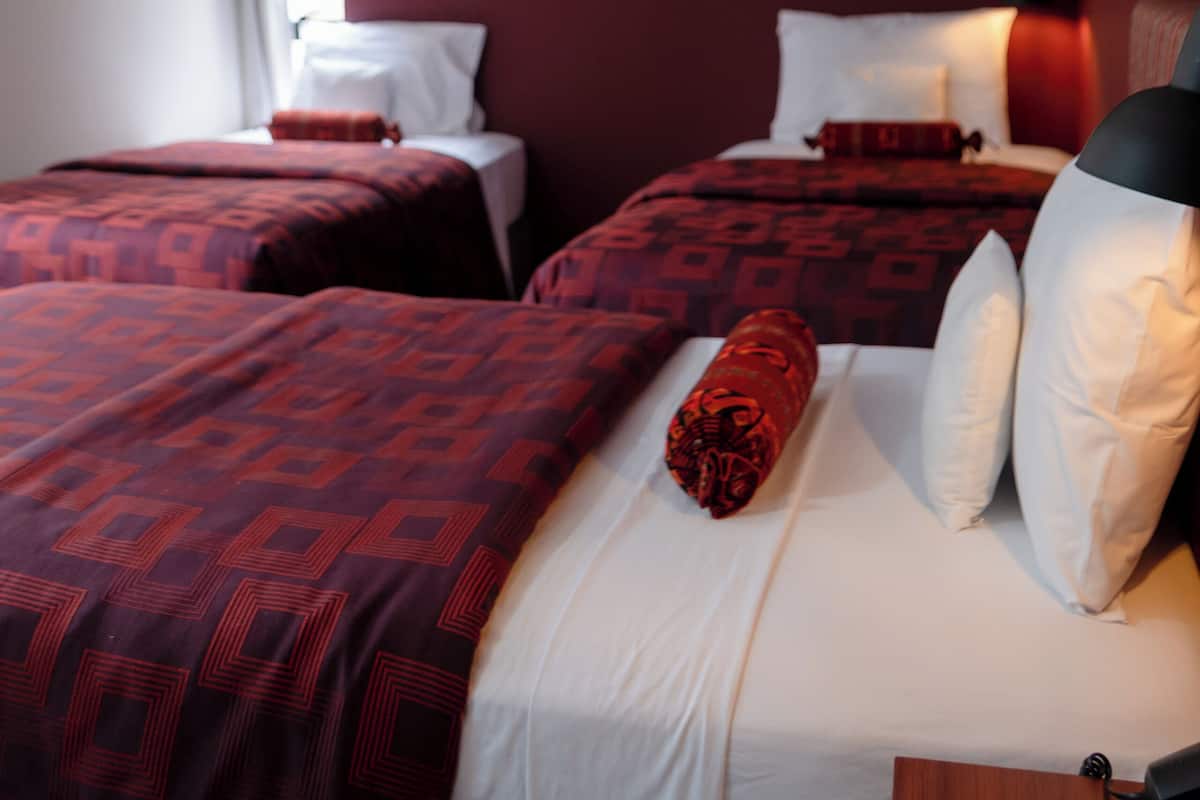 Ein Hotelzimmer mit zwei Betten, jeweils mit rot gemusterten Bettbezügen und weißen Kissen. Links befindet sich ein Fenster, ein an der Wand befestigtes Fernsehgerät und ein Nachttisch mit einer Lampe.