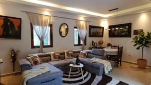 A living room with a sectional sofa, a small round table, a dining table with chairs, and a potted plant.