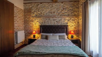 A bedroom with a stone wall, a wooden ceiling, a bed with a patterned bedspread, two bedside lamps, and a window with curtains.