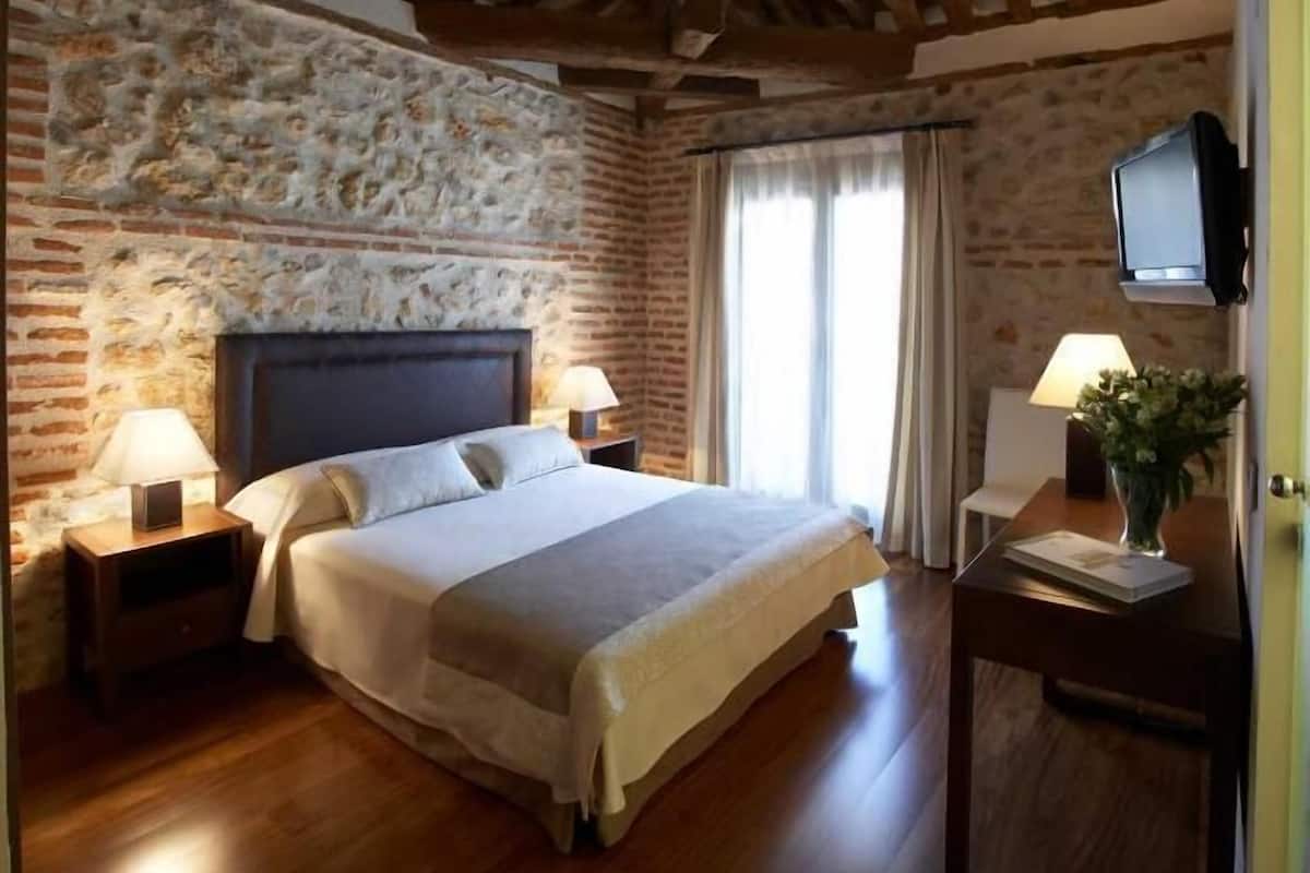 A bedroom with a stone wall, wooden beams, a bed, two bedside tables with lamps, a TV on a stand, and a window with curtains.