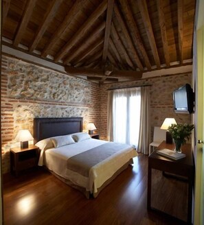 Un dormitori amb paret de pedra, bigues de fusta, un llit, dues tauletes de nit amb làmpades, un televisor sobre un suport i una finestra amb cortines.
