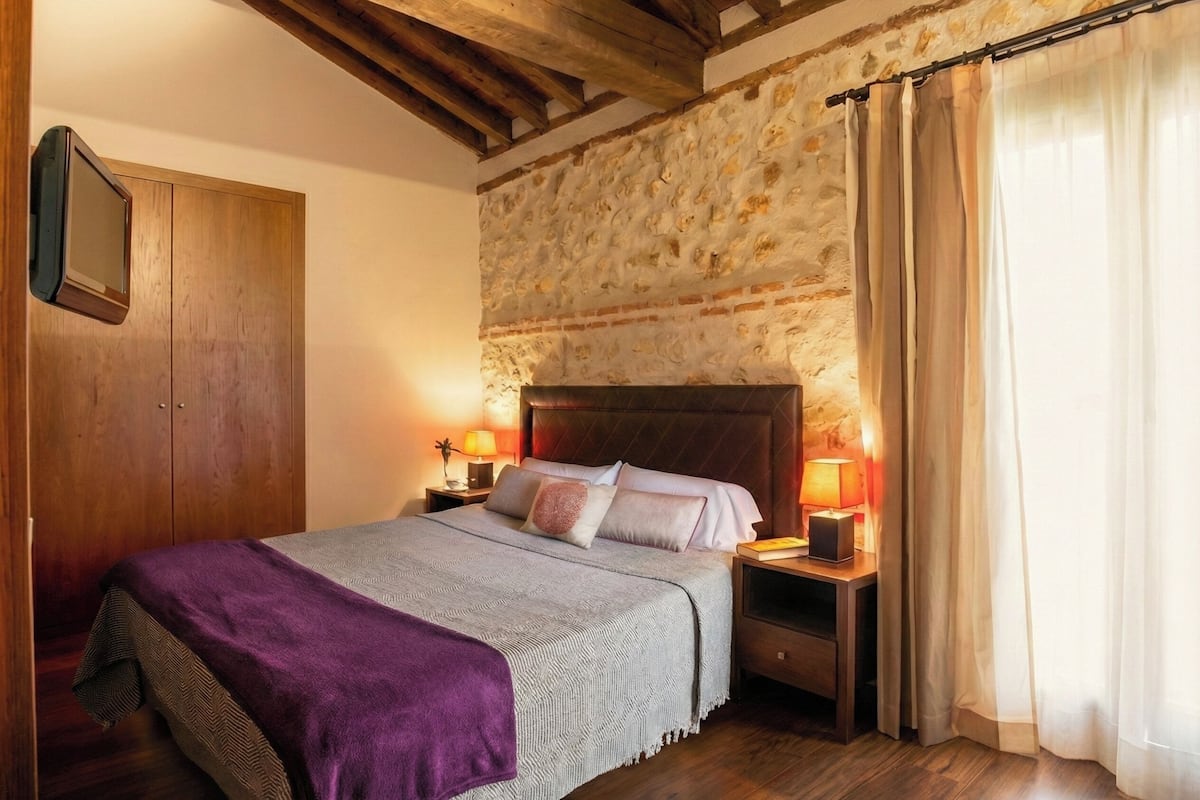 A bedroom with a stone wall, wooden ceiling beams, a bed with a purple blanket, two bedside lamps, and a wooden wardrobe.
