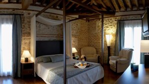 Un lit à baldaquin dans une chambre rustique aux murs de pierre, au plafond en bois et agrémentée de deux fauteuils.