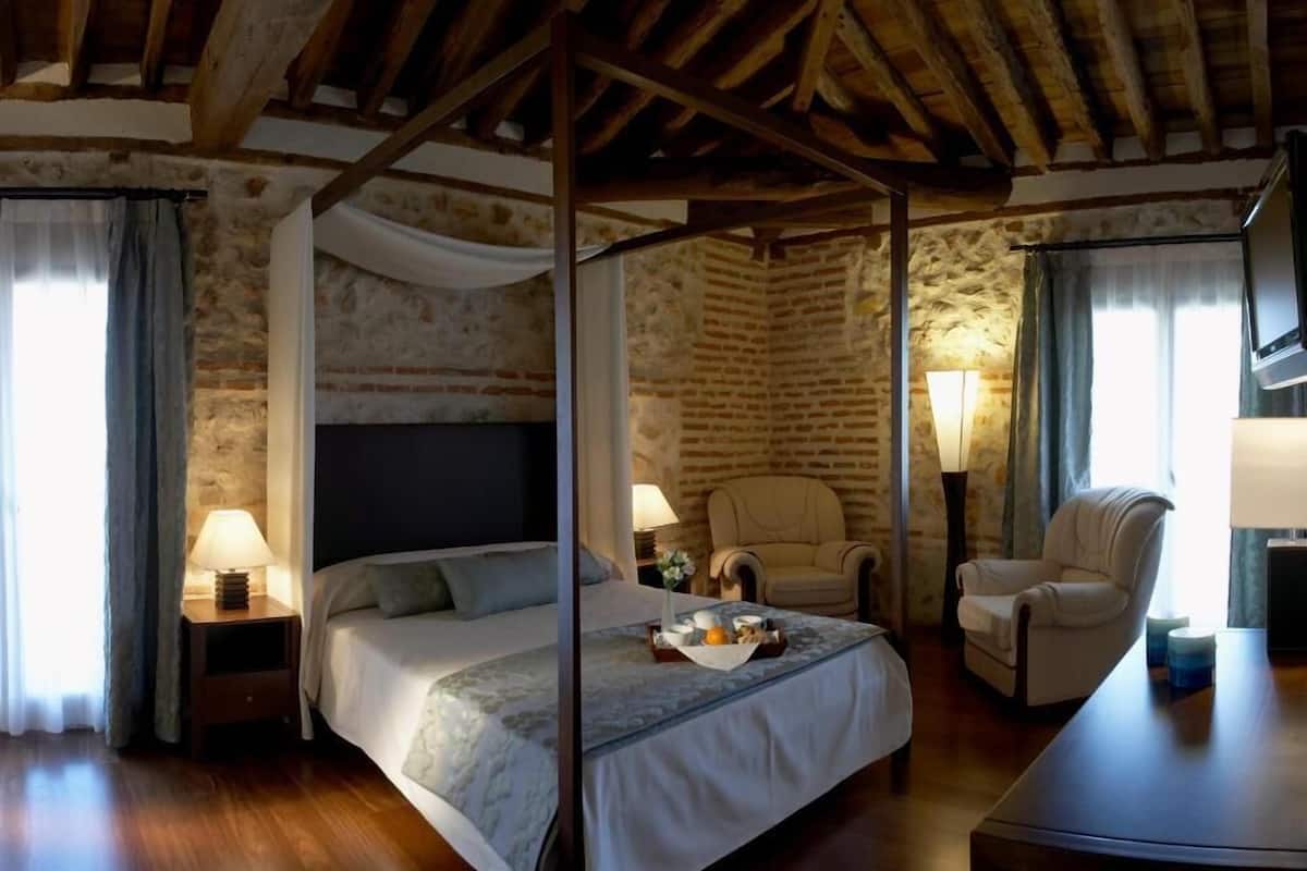 A four-poster bed in a rustic room with stone walls, a wooden ceiling, and two armchairs.