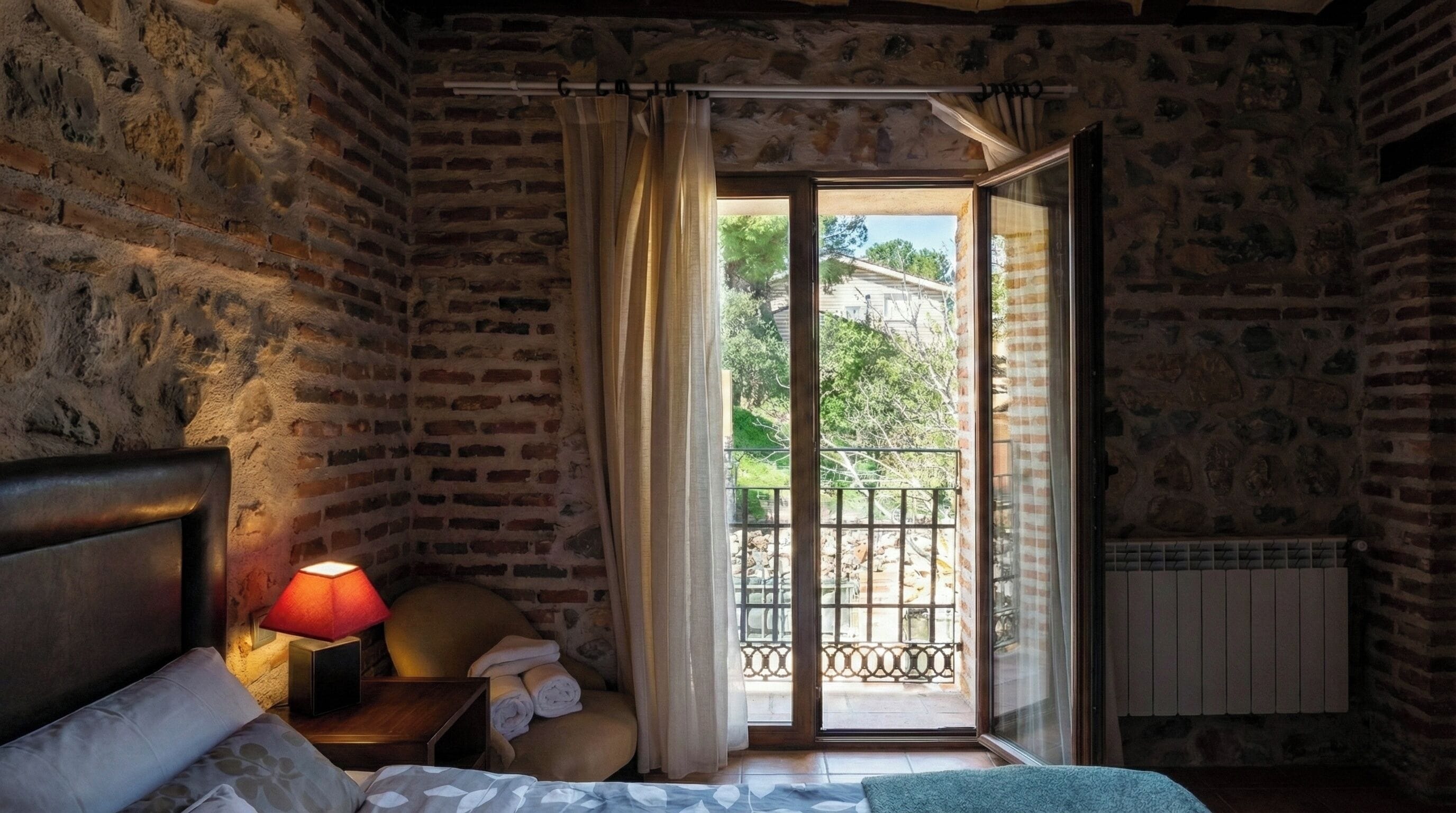 Quarto com cama, mesa de cabeceira com abajur, cadeira e vista para a varanda.