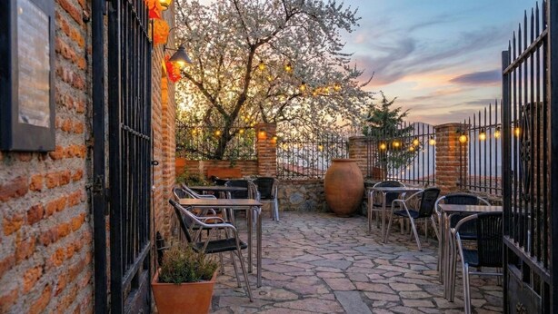 Une terrasse extérieure avec un sol en pierre, des chaises et des tables en métal, entourée de murs en briques et d’une clôture en métal.