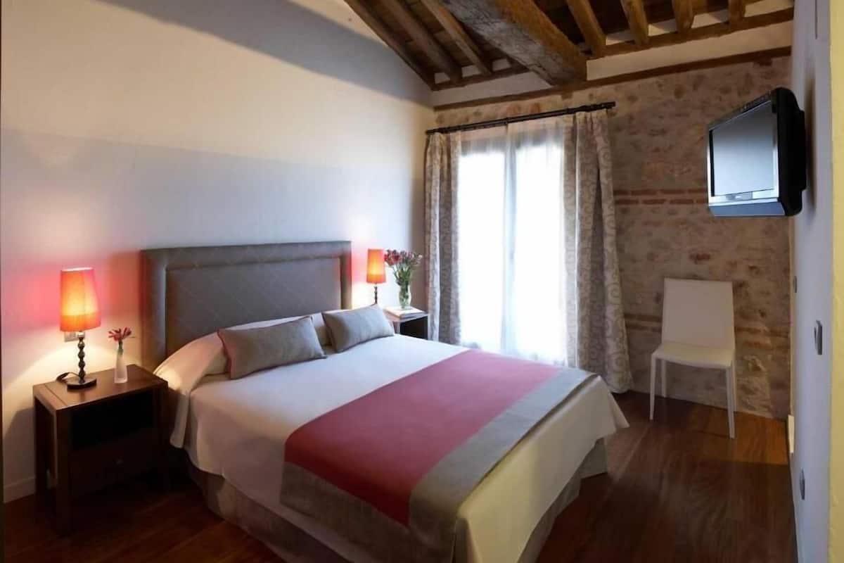A bedroom with a bed, nightstands, a TV, and a window with curtains.