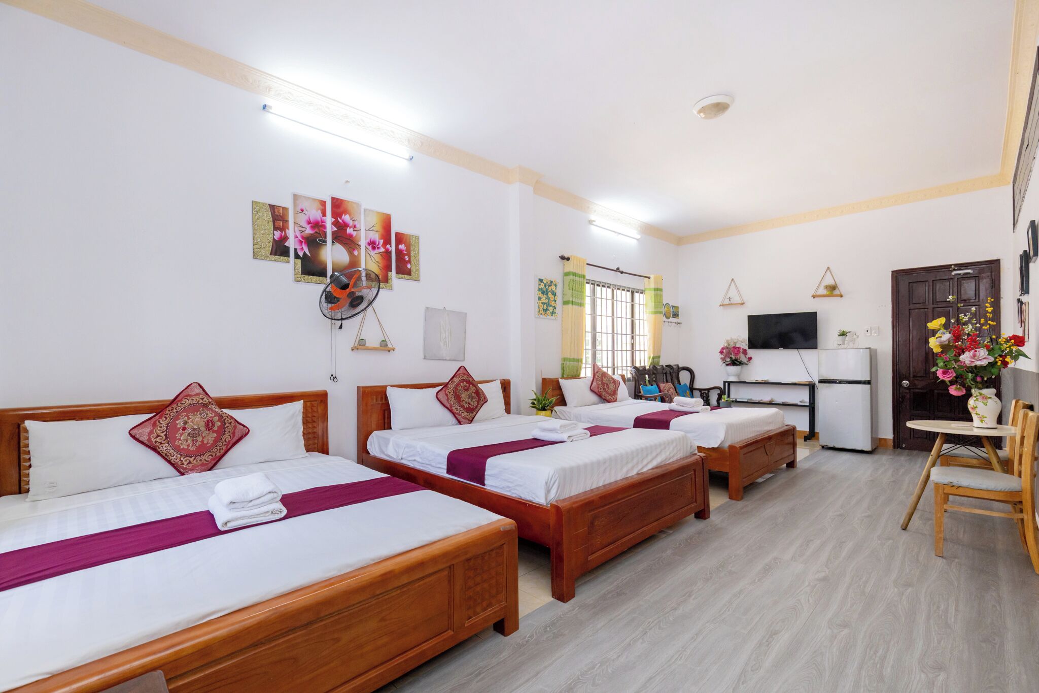 Une chambre d’hôtel avec trois lits en bois, une télévision, un réfrigérateur et un coin repas comprenant une table et des chaises.