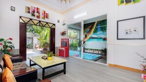 Un hall de hotel con un sofá, una mesa y una máquina recreativa roja. Se observa un ventanal con un mural tropical y vista a una piscina.