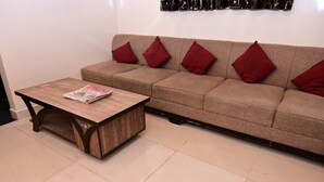 A living room with a wooden coffee table, a beige sofa with red cushions, and a tiled floor. - Hotel Vinayak Near Akshaya Patra Jagatpura (Jaipur)