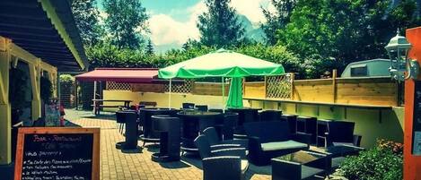 An outdoor café with tables and chairs, a green umbrella, and a signboard with menu items.