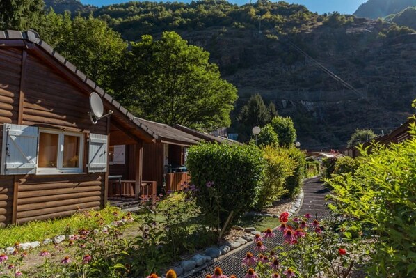 A wooden cabin with a balcony, surrounded by lush greenery and a mountain backdrop. - ZEcamping 4* piscine chauffée club enfant locatif TV BBQ 35m2 5 pers (Le Bourg-d'Oisans)