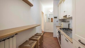 A narrow kitchen with white cabinets, a stove, and a microwave. There are two stools and a wooden shelf. - Old City Nest by MMega (Florence)